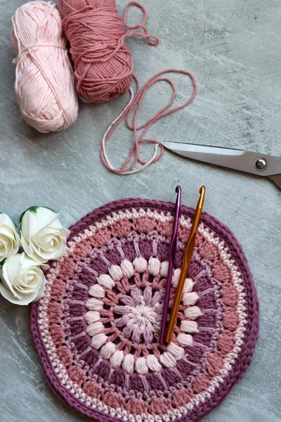 Crochet hook with cotton thread, scissors and roses on grey table, closeup photo of handmade pattern. Leisure and hobby concept. Space for text.