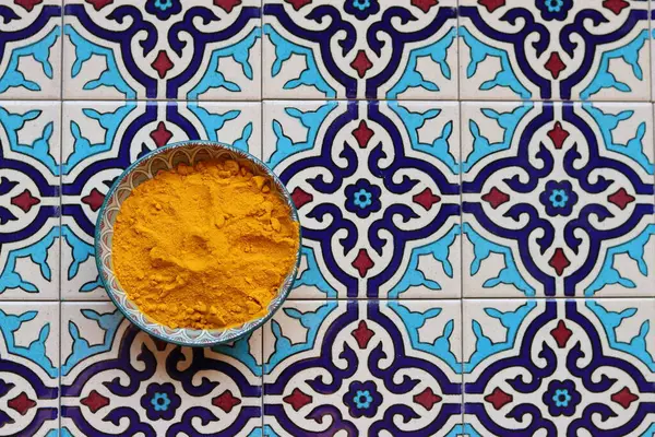 Turmeric powder in a ceramic bowl on Moroccan tiled blue background with copy space. 