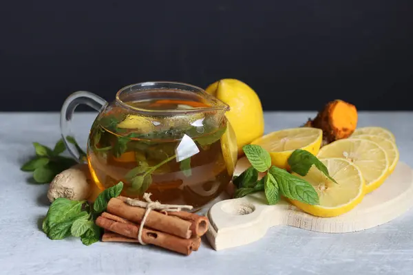 Teapot with hot tea, ginger, turmeric root, cinnamon sticks, lemon and mint leaves on grey background with copy space. Anti inflammatory winter drink recipe.