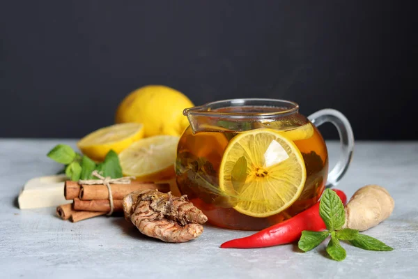 Teapot with hot tea, ginger, turmeric root, cinnamon sticks, lemon and mint leaves on grey background with copy space. Anti inflammatory winter drink recipe.