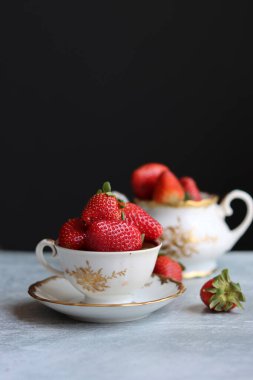 Küçük beyaz ve altın porselen bir fincanda çilekler koyu arka planda ve metin için yer var. İçinde retro sofra takımı ve yaz meyveleri olan natürmort fotoğraf. Taze konsept yemek.