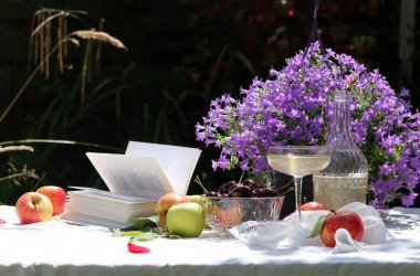 Açık bir kitap, kiraz, elma, şarap kadehi ve bahçe masasında Campanula çiçekleri olan bir hayat. Piknik masasının parlak resmi. Yaz tatili aktivite kavramı. 