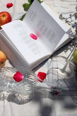 Açık bir kitap, gül yaprakları, elmalar, kirazlar ve beyaz bir masa örtüsü üzerinde şarap. Bahçe masasında yaz hala hayattadır. Boş zaman aktiviteleri konsepti. 