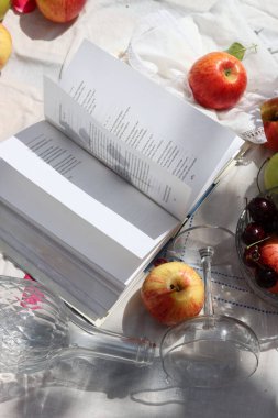 Açık bir kitap, gül yaprakları, elmalar, kirazlar ve beyaz bir masa örtüsü üzerinde şarap. Bahçe masasında yaz hala hayattadır. Boş zaman aktiviteleri konsepti. 