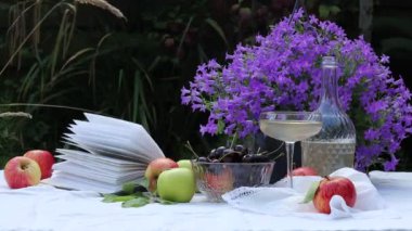 Bahçede piknik. Güneşli bir yaz günü. Tencerede güzel mavi Campanula çiçekleri, kitabın sayfaları rüzgârla savruluyor. Beyaz masa örtüsü üzerinde taze organik meyve ve gül yaprakları.. 