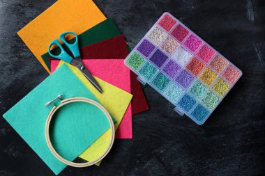 DIY projesi yapılıyor. Renkli cam boncukların en üstteki fotoğrafı, koyu gri masada kumaş ve makas. İşlem sırasında el işi. 