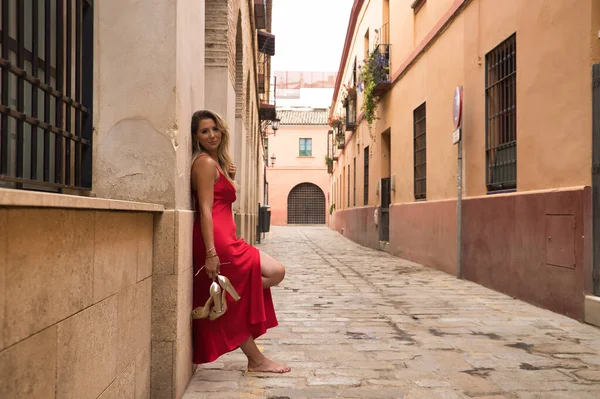 stock image Young, attractive, blonde woman, wearing an elegant red party dress and holding golden high heels in her hand, barefoot, leaning against the wall of a city alley. Concept beauty, fashion, elegance.