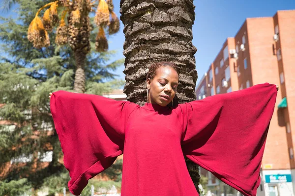 身穿漂亮红色派对礼服 袖子很大的非洲裔美国女人靠在棕榈树树干上摆姿势 概念时尚 — 图库照片