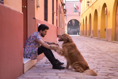 Kapıda oturan sakallı genç İspanyol adam köpeğinin patisini sevgiyle tutuyor. Hayvanlar, köpekler, aşk, evcil hayvanlar, altın.