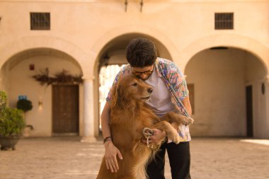 Sakallı ve güneş gözlüklü genç İspanyol adam köpeğini çok mutlu bir şekilde kucaklıyor. Hayvanlar, köpekler, aşk, evcil hayvanlar, altın.