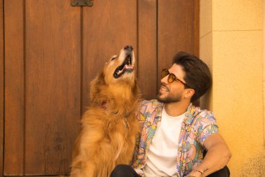Young Hispanic man with beard and sunglasses sitting next to his dog on the front step of a house, very happy. Concept animals, dogs, love, pets, golden.