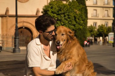Young Hispanic man with beard, sunglasses and white shirt, sitting on a bench hugging and petting his dog. Concept animals, dogs, love, pets, golden.