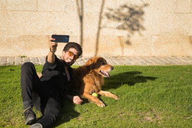 Sakallı, güneş gözlüklü, siyah gömlekli, cep telefonuyla selfie çeken, köpeğinin yanında güneşlenen genç bir İspanyol. Hayvanlar, köpekler, aşk, evcil hayvanlar, altın.