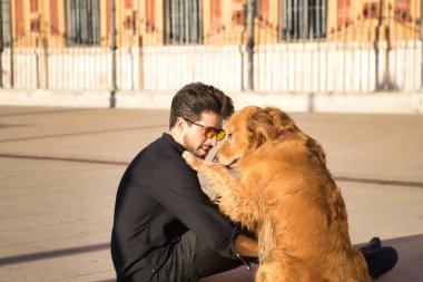 Sakallı, güneş gözlüklü, siyah gömlekli, yerde köpeğiyle oturan çok sevecen ve suç ortağı bir genç. Hayvanlar, köpekler, aşk, evcil hayvanlar, altın.