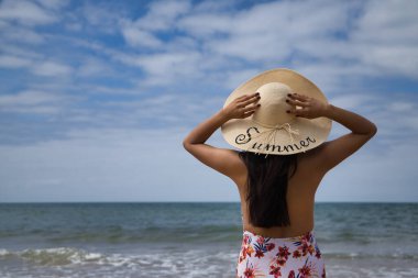 South American woman, young and beautiful, brunette with hat with the word summer held with both hands and swimsuit on her back looking at the sea. Concept sea, sand, sun, beach, vacation, travel.