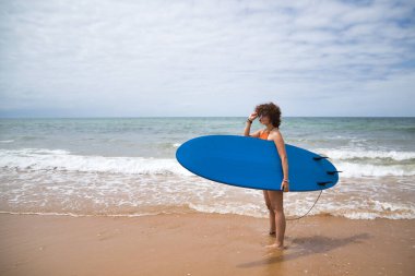 Attractive mature woman with curly hair, sunglasses and bikini, posing holding a blue surfboard under her arm. Concept sea, sand, sun, beach, vacation, surf, summer.