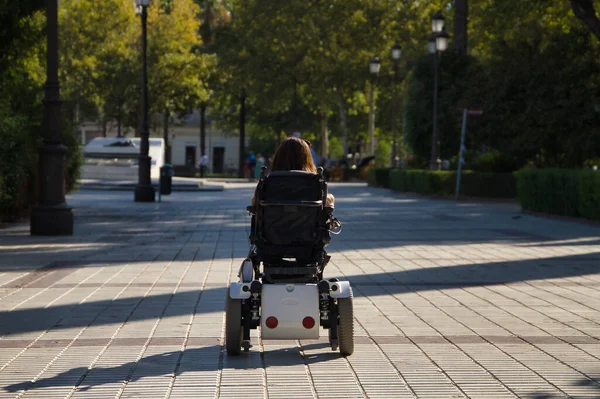 Woman with disability, reduced mobility and small stature in an electric wheelchair walking through the city center, seen from the back. Concept handicap, disability, incapacity, special needs.