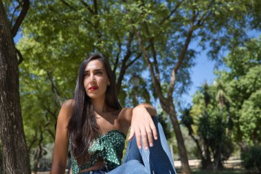 Genç bir kadın, esmer, ince, yeşil tişört ve kot giymiş, taş bir bankta oturuyor, yalnız, üzgün, ciddi. Yalnızlığı, kederi, üzüntüyü kavra..