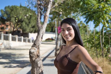 Güney Amerikalı bir kadın, genç ve güzel, esmer, kahverengi elbiseli, kulağında güzel bir çiçekle korkuluklara yaslanmış. Moda, güzellik, çiçekler, seyahat, tatiller, rahatlama.