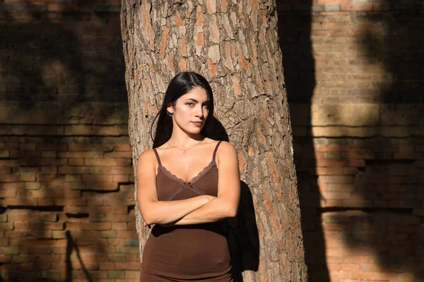 南アメリカの女性は 若くて美しい ブルネット 茶色のドレスで 彼女の腕を持つ木の幹にもたれて 怒って渡った 概念の怒り 深刻さ 嫌悪感 — ストック写真