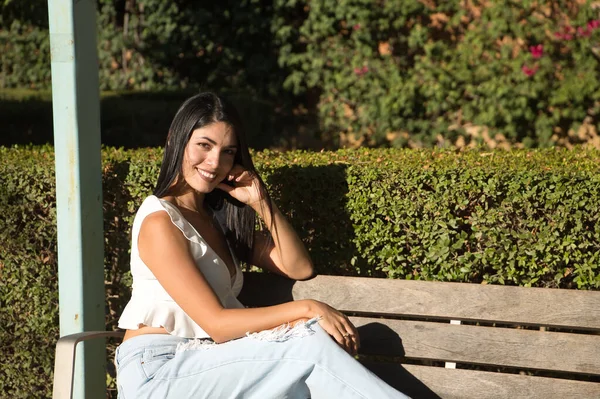 南アメリカの女性は 若くて美しい ブルネット 白いシャツとジーンズを着て 木製のベンチに座って 笑顔と幸せ コンセプトの美しさ ファッション トレンド — ストック写真