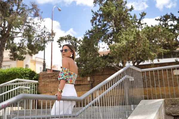 Çiçekli gömlekli, beyaz pantolonlu, güneş gözlüklü, Avrupa 'da tatilde korkuluklara dayanan genç, çekici, sarışın bir kadın. Güzelliği, modayı, zarafeti, seyahati kavra.