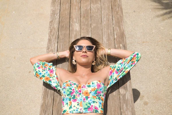 Genç, çekici, sarışın bir kadının portresi. Çiçekli gömlek ve güneş gözlüğü takıyor. Elleri başının arkasında, tahta bir bankta yatıyor. Güzellik, moda, trend, rahatlama, tatil, seyahat kavramı.