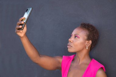 Pembe tişörtlü ve pembe göz falı Afro-Amerikan kadın portresi gri arka planda akıllı telefonuyla selfie çekiyor. Mobil konsept, fotoğraflar, selfie, sosyal ağlar.