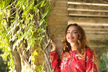 Altın ve gümüş ipliklerle işlenmiş tipik bir Fas kırmızı elbisesi içindeki genç ve güzel bir kadın, sarmaşıklı tuğla bir sütunun yanında poz veriyor. Güzellik kavramı, etnik köken, tipik takım elbiseler, Marakeş, Arap.