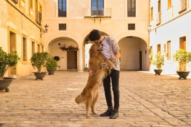 Sakallı ve güneş gözlüklü genç İspanyol adam köpeğini çok mutlu bir şekilde kucaklıyor. Hayvanlar, köpekler, aşk, evcil hayvanlar, altın.
