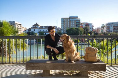 A young Hispanic man with a beard, sunglasses, black shirt and backpack, climbed on a bench with his dog while petting him under the sun's rays. Concept animals, dogs, love, pets, golden.