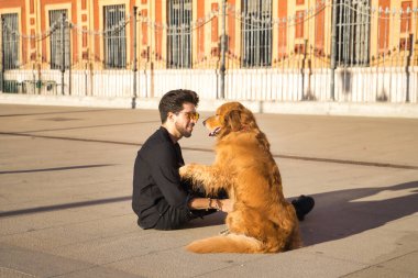 Sakallı, güneş gözlüklü, siyah gömlekli, yerde köpeğiyle oturan çok sevecen ve suç ortağı bir genç. Hayvanlar, köpekler, aşk, evcil hayvanlar, altın.