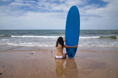 South American woman, young and beautiful, brunette with sunglasses and bikini, sitting on her back and holding a blue surfboard. Concept sea, sand, sun, beach, vacation, surf, summer.