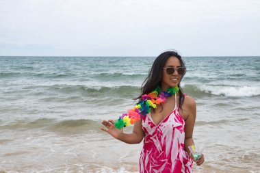 Young and beautiful South American woman, brunette, with sunglasses, bikini and flower necklace with a glass of blue wine in her hand, very happy and smiling. Summer concept, beach, drinks.