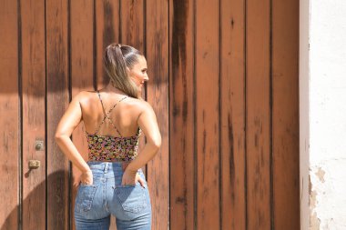 Genç ve güzel bir kadın, at kuyruklu, çiçekli bir bluz ve kot pantolon giyen, arkası dönük ve elleri pantolonunun arka cebinde sıkışmış. Güzelliği, modayı, trendi, kotu kavra.