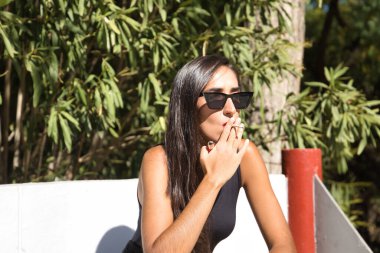 Genç bir kadın, esmer, ince, siyah giyinmiş, güneş gözlüğü takıyor, bankta oturmuş sigara içiyor. Kavram tütün, sigara, bağımlılık, sağlık, sigara.