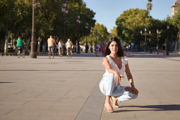 South American woman, young and beautiful, brunette, with white shirt and jeans, crouching and posing happily looking at the camera. Concept beauty, fashion, trend, model.