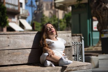 Özürlü bir kadın, hareket kabiliyeti düşük ve ufak tefek, tahta bir bankta oturuyor, mutlu ve rahat bir şekilde güneşleniyor. Özürlülük, yetersizlik, yetersizlik, özel ihtiyaçlar, üstesinden gelmek.