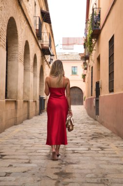 Genç sarışın kadın, arkası dönük, zarif kırmızı bir parti elbisesi giyiyor ve elinde altın topuklularla şehir sokaklarında yalınayak yürüyor. Güzelliği, modayı, zarafeti, dikey.