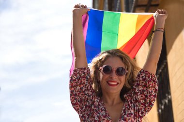 Orta yaşlı sarışın bir kadın, çiçekli bir elbise ve güneş gözlüğü takıyor, sokakta yürüyor, el sallıyor ve eşcinsel gurur bayrağı sallıyor. Kavram lgtbi, gay, lezbiyen, gurur günü.