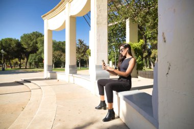 Genç bir kadın, esmer, ince, siyah giyinmiş ve güneş gözlüğü takıyor, cep telefonunda sosyal ağlara danışmanlık yapıyor. Güzelliği, modayı, uygulamaları, interneti, akıllı telefonu kavra.