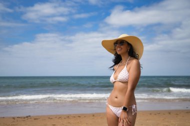 South American woman, young and beautiful, brunette with sunglasses, hat and bikini posing happy and smiling on the beach. Concept sea, sand, sun, beach, vacation, travel, summer.