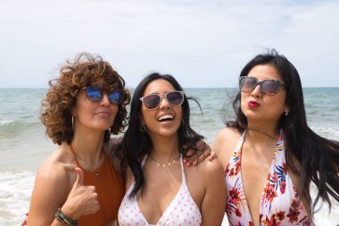 Attractive mature woman next to two young, pretty, brunette South American women in bikinis and sunglasses taking funny pictures of each other. Concept vacation, friends, multiethnic, summer.