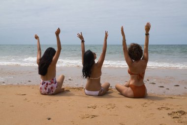 Çekici olgun kadın, bikinili ve güneş gözlüklü, kolları havada kumda oturan iki genç, güzel, esmer Güney Amerikalı kadının yanında mutlu. Tatil kavramı, arkadaşlar, çok ırklı yaz..