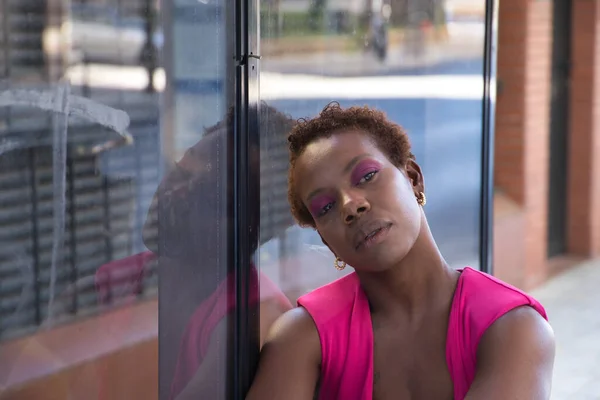 Portrait of African American woman in a pink T-shirt with her head leaning against the glass of a bus stop with distracted look. Concept transportation, thoughts, distraction, waiting.