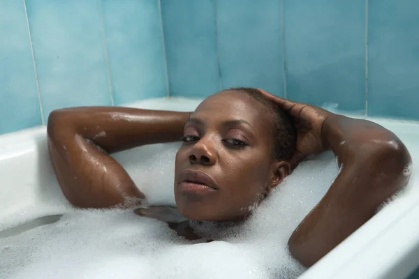 Portrait of African American woman, with her hands on her head and taking a bath with the bathtub full of foam, very relaxed. Concept bath, relax, foam, soap.