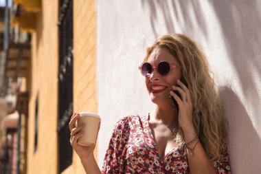 Orta yaşlı sarışın kadın, çiçekli bir elbise ve güneş gözlüğü takıyor elinde bir bardak kahveyle cep telefonunda gülümsüyor. Kavram iletişimi, cep telefonu, konuşma.