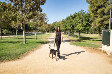 Genç bir kadın, esmer, zayıf, siyah giyinmiş, güneş gözlüğü takıyor, köpeğini parkta gezdiriyor. Güzelliği, modayı, hayvanları, hayvanları, melezi kavra.