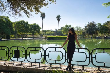 Genç bir kadın, esmer, ince, siyah giyinmiş ve güneş gözlüğü takıyor, yeşil renkli bir gölün yanındaki korkuluklara yaslanıyor. Güzelliği, modayı, suyu, açık havayı kavra.