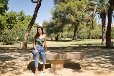 Genç bir kadın, esmer, ince, yeşil tişört ve kot giymiş, taş bir bankta oturuyor, yalnız, üzgün, ciddi. Yalnızlığı, kederi, üzüntüyü kavra..